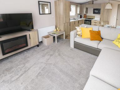 A living room with a large gray sofa with yellow and orange cushions a TV on a stand with a fireplace below a small table with candles and magazines and a kitchen area with wooden cabinets at The Waves Little Haven near Broad Haven