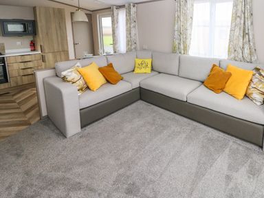 A living area with a sectional sofa with colorful cushions near windows with patterned curtains and a kitchen with wooden cabinets at The Waves in Little Haven near Broad Haven