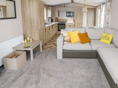 A living room with a grey carpet sectional sofa with yellow pillows a beige side table and a kitchen with wooden cabinets at The Waves in Little Haven near Broad Haven