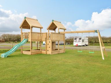 A playground with wooden climbing structures slides and swings on grass with a caravan in the background at The Waves in Little Haven near Broad Haven