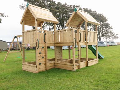 A wooden outdoor playground structure with two towers connected by a bridge a slide a climbing wall and swings on a grassy area at The Waves in Little Haven near Broad Haven