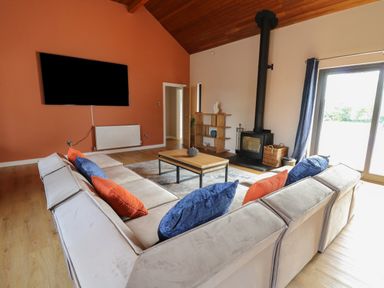 A living room with a beige sectional sofa with blue and orange pillows a wooden coffee table a wall-mounted TV a wood-burning stove and a wooden ceiling at Heritage in St Clears