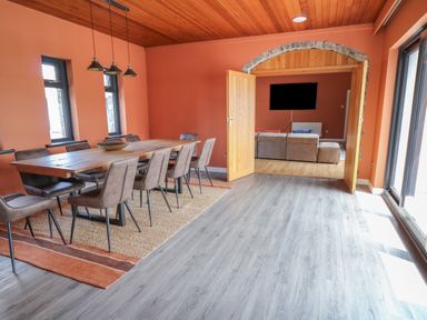 A dining room with a wooden table and eight chairs a rug under the table wooden ceiling and orange walls double doors open to a living room with a sofa and a wall mounted TV at Heritage St Clears
