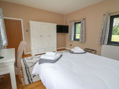 A bedroom with a bed with folded towels a white wardrobe a wall-mounted TV two windows with patterned curtains and a chair at Heritage in St Clears