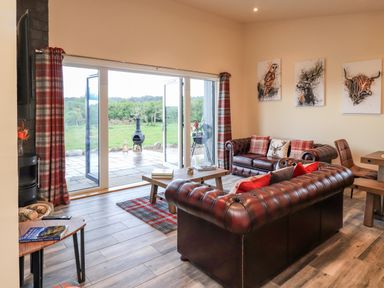 A living room with leather sofas and plaid curtains facing glass doors overlooking a patio and green landscape at Pine Marten in Dornoch