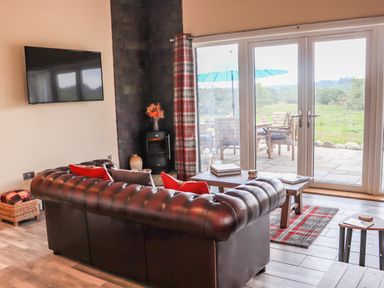 A living room with a leather sofa a wall-mounted tv a coffee table and glass doors opening to a patio at Pine Marten in Dornoch
