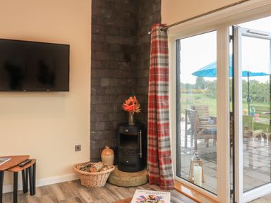 A corner of a living room with a wall-mounted television a small stove with flowers on top a basket of firewood and a sliding glass door with patio furniture outside at Pine Marten in Dornoch