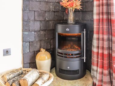 A black electric stove with a glass vase of orange leaves on top a basket of firewood and a brown jug near a red and gray plaid curtain at Pine Marten in Dornoch
