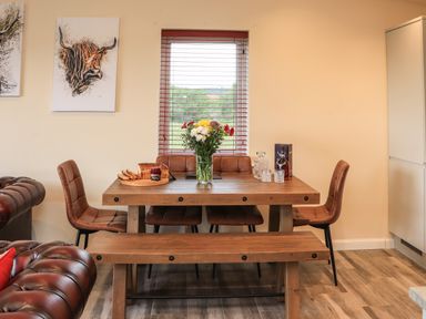 A dining area with a wooden table, leather chairs, a bench, flowers in a vase, bread and jam on a tray, and glassware beside a window with wooden blinds at Pine Marten in Dornoch