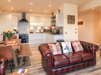 A kitchen with white cabinets and countertop next to a wooden dining table with flowers on it and a brown leather sofa with cushions at Pine Marten in Dornoch