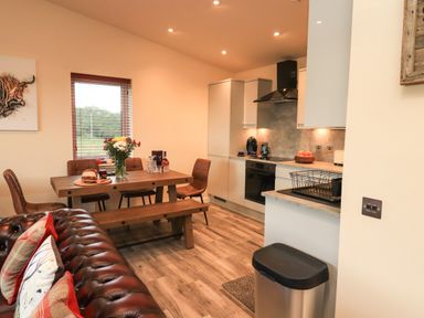 A kitchen and dining area with a wooden table, brown chairs, a vase of flowers, kitchen appliances, and a brown leather sofa at Pine Marten in Dornoch