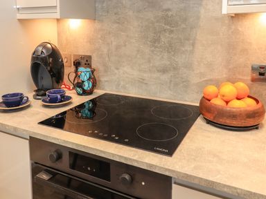 A kitchen countertop with a black stove two blue cups with saucers a coffee pod holder a coffee machine and a wooden bowl with oranges at Pine Marten in Dornoch
