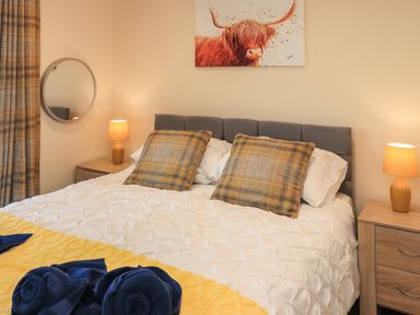 A bedroom with a bed with white bedding and plaid pillows two bedside tables with lamps and a round mirror above one table at Pine Marten in Dornoch