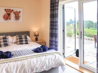 A bedroom with a bed featuring blue plaid pillows and rolled towels next to glass doors with plaid curtains at Pine Marten in Dornoch