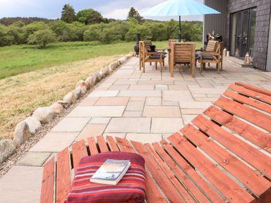 An outdoor patio with wooden chairs and a table under a blue umbrella next to a stone paved area with a wooden bench holding a striped blanket and book at Pine Marten in Dornoch