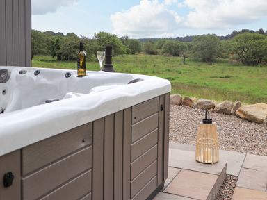 An outdoor hot tub with a bottle and glass on its edge overlooking a grassy field with trees and rocks near Pine Marten in Dornoch