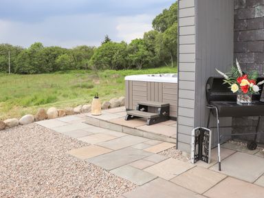 An outdoor patio with a hot tub, small steps, a barbecue grill with a flower vase, a tool kit, and a lantern by a grassy field at Pine Marten in Dornoch