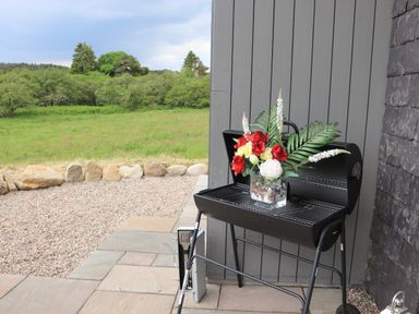 A barbecue grill with a vase of flowers on a patio next to a gray wall with a grassy field in the background at Pine Marten in Dornoch