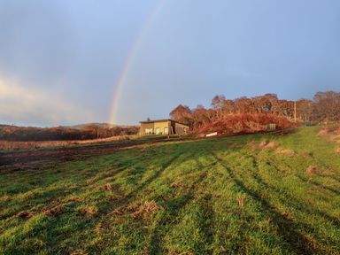 A grassy field with tire tracks leading to a modern house and a rainbow in the sky at Pine Marten in Dornoch