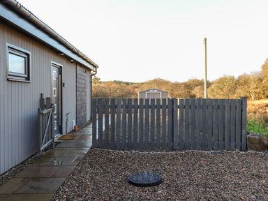 A gray exterior house wall with a door and window next to a gray wooden fence and a gravel yard with a small shed in the background at Pine Marten in Dornoch