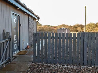 A gray wooden fence and gate in front of a house with a gray door and a shed in the background at Pine Marten in Dornoch