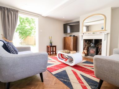 A living room with two gray armchairs a white coffee table with chess and other games a Union Jack rug a fireplace a wall-mounted TV and a small plant at Cavan Cottage in Yoxford