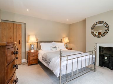 A bedroom with a metal bed frame bedside tables with lamps a wooden dresser a round mirror above a fireplace and a wooden door at Cavan Cottage in Yoxford