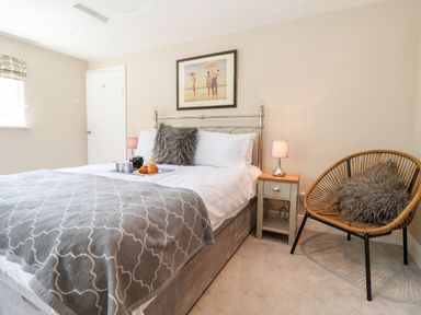 A bedroom with a double bed bedside tables lamps a wicker chair with a cushion and a painting on the wall at Cavan Cottage in Yoxford