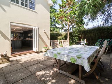 An outdoor patio with a table covered by a tablecloth surrounded by wooden chairs next to an open door leading to a kitchen at Cavan Cottage in Yoxford
