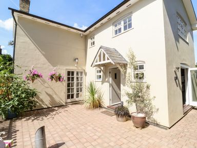 A paved courtyard with potted plants hanging flowers and a cream house with a doorway and windows at Cavan Cottage in Yoxford