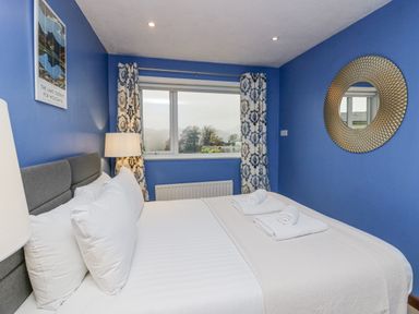 A bedroom with a made bed with white linens pillows and towels on it a window with patterned curtains a bedside lamp and a round mirror on the wall at Fairfield View in Ambleside