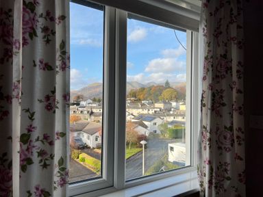 A view through a window with floral curtains showing houses trees and mountains at Fairfield View in Ambleside