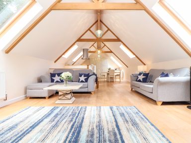 A living area with two grey sofas, a wooden coffee table with flowers, a striped blue rug, and a dining table with chairs at Potters Barn in Trevarno near Helston