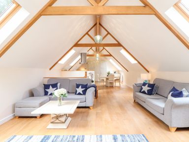 A loft living area with grey sofas, star patterned cushions, a coffee table, wood beams and a dining table at Potters Barn Trevarno near Helston