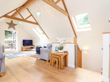 A living area with wooden floor and beams two gray armchairs a wooden TV stand with television and doors leading outside at Potters Barn in Trevarno near Helston