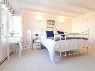 A bedroom with a metal bed frame, two navy pillows, a mirrored bedside table with a lamp, a white desk with chair, and a window at Potters Barn Trevarno near Helston