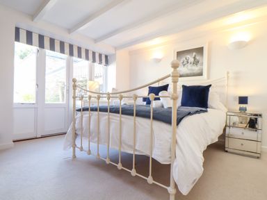 A bedroom with a white metal bed frame blue pillows white bedding striped window blinds and a mirrored bedside table at Potters Barn Trevarno near Helston