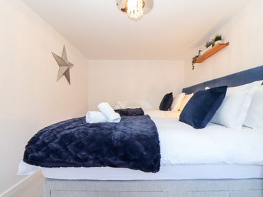 A bedroom with a bed covered in white linens and navy blue blankets and pillows with a metal star decoration on the wall and a wooden shelf with plants at Potters Barn Trevarno near Helston