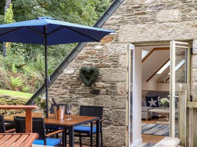 An outdoor patio area with a wooden table, blue chairs, and a blue umbrella next to a stone wall and open double doors leading to a living room with a gray couch and a star pillow at Potters Barn in Trevarno near Helston