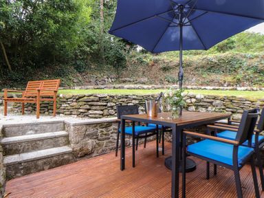 An outdoor patio with a table and four chairs under a blue umbrella next to stone steps and garden walls at Potters Barn Trevarno near Helston