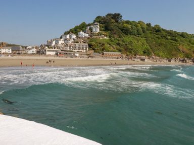 A beach with waves and people walking near buildings at the base of a green hill at 3 River View in Looe