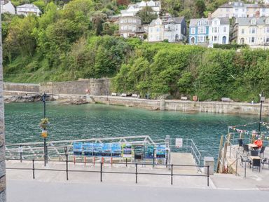 A waterfront with a pier and seating area overlooking green water and hillside houses at 3 River View in Looe