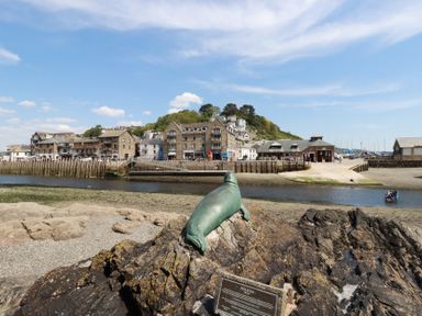 A bronze seal sculpture on rocks overlooking a river with stone buildings and green hillside in the background at 3 River View in Looe