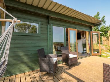 An outdoor wooden deck with two wicker chairs and a hammock outside a green wooden cabin with glass doors open at Dart View Hideout in Totnes