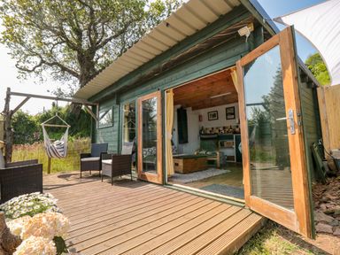 An outdoor wooden deck with chairs and a hanging chair next to a small cabin with open wooden framed glass doors at Dart View Hideout in Totnes