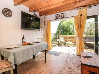 A dining table with plates wine and bread under a wall mounted television in a room with wooden ceiling beams and an open door to a deck with chairs and plants at Dart View Hideout in Totnes