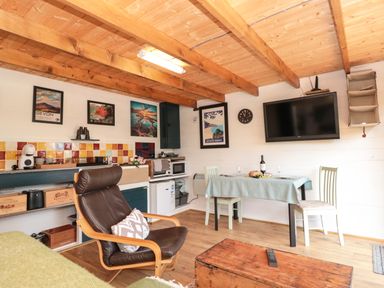 A small living area with a brown leather chair a table set for two under a wall-mounted TV and kitchen appliances on shelves at Dart View Hideout in Totnes