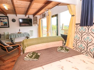 A room with two beds, a dining table with two chairs, a mounted TV, and large windows with yellow curtains at Dart View Hideout in Totnes