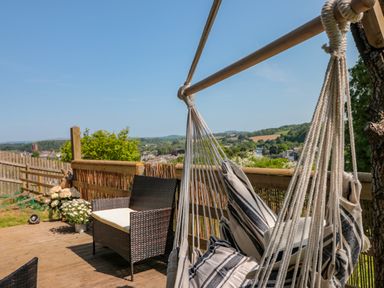 An outdoor deck with a hanging chair with striped cushions a wicker loveseat and potted flowers overlooking a landscape at Dart View Hideout in Totnes