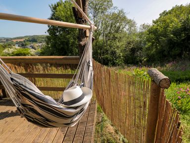 A striped hammock with a white hat on a wooden deck overlooking a garden with trees at Dart View Hideout in Totnes